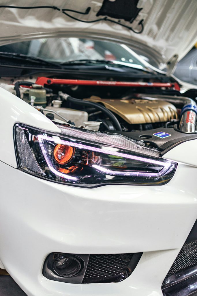 Close-up of a car's engine and headlight under the open hood in a garage.
