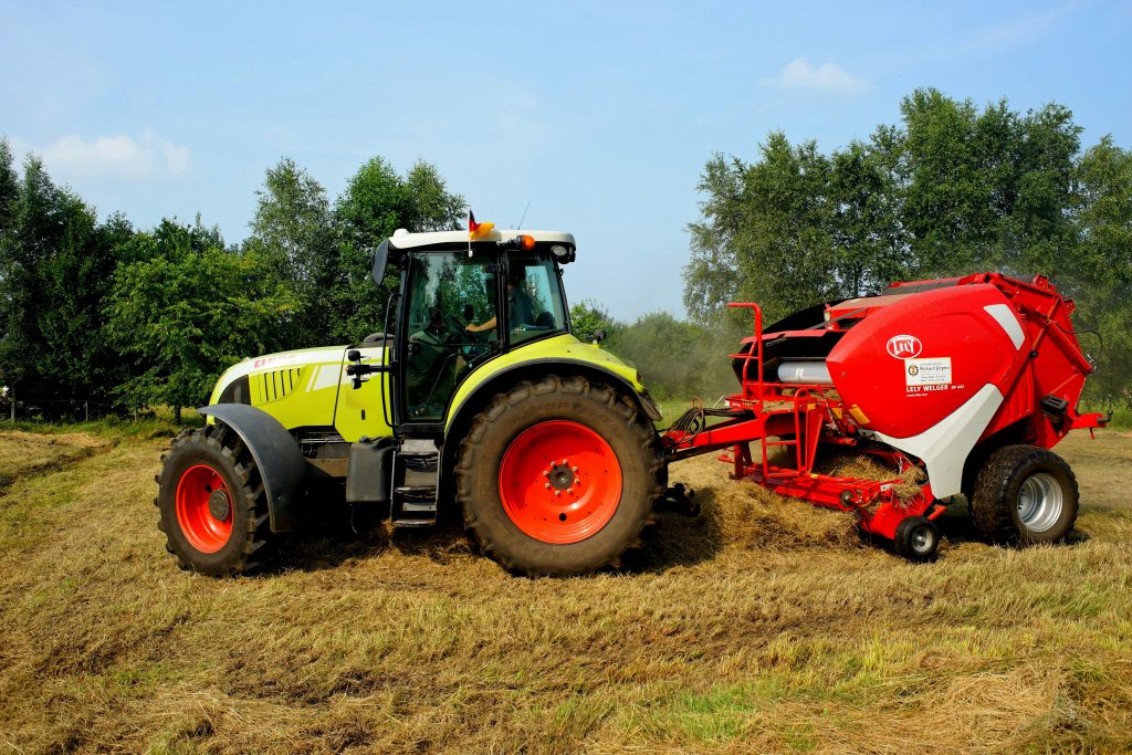 Green tractor and hay baler working on a farm field with trees in the background.